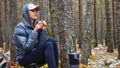 Woman hiker in jacket drinks hot tea in forest camping sitting on chair.