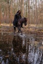 A woman in a historical costume with a bow and arrows riding a horse through a forest swamp