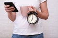 Woman holding a big pink cup of coffee and alarm clock showing 7 am in hands, she using a smartphone and solving business cases Royalty Free Stock Photo