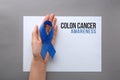 Woman holding blue ribbon near card with words COLON CANCER AWARENESS on grey background, top view