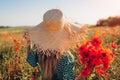 Woman holding bouquet of poppies flowers walking in summer field. Stylish girl in straw hat enjoys landscape