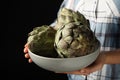 Woman holding bowl with fresh raw artichokes on black background, closeup Royalty Free Stock Photo