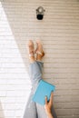 Woman holding an close book with her legs up in blue jeans and pink sneakers against a brick wall