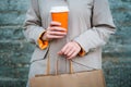 Woman holding coffee in disposable orange cup and paper shopping bag.