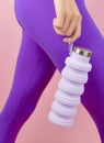 Woman holding collapsible reusable lilac water bottle on pink background. Sustainable lifestyle