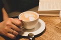 woman holding cup of coffee with milk foam near book on wooden table in cafe Royalty Free Stock Photo