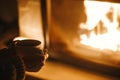 Woman holding cup of tea and warming up hands at cozy fireplace in dark evening room, close up. Fireplace heating in house,