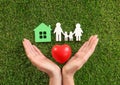 Woman holding hands near red heart and figures house and family on green grass, top view