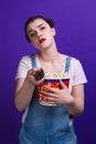 Woman holding large popcorn box, staring change channel while watching television with remote control isolated at purple Royalty Free Stock Photo