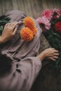 Woman holding orange dahlia flower and sitting on wooden rustic bench, view above. Atmospheric moody image. Florist in linen dress