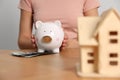 Woman holding piggy bank, house model and banknotes at wooden table, closeup Royalty Free Stock Photo