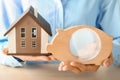 Woman holding piggy bank with house model at wooden table, closeup Royalty Free Stock Photo