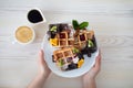 Woman holding plate with belgian waffles decorated with fruits and syrup, sitting at table with cup of coffee, top view