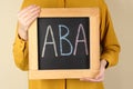 Woman holding small chalkboard with abbreviation ABA Applied behavior analysis on beige background, closeup