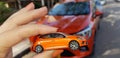 Woman holding small orange car model against real car of same type