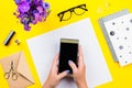 Woman holding a smartphone over a bright yellow desktop with documents, accessories and flowers layout Royalty Free Stock Photo