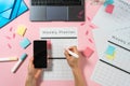 Woman holding smartphone and writing at planner on pink pastel background with laptop and stationery