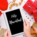 Woman holding tablet with text Feliz Navidad in Spanish language that means Merry Christmas, red poinsettia flower, gift boxes on Royalty Free Stock Photo