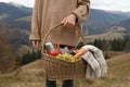 Woman holding wicker picnic basket with thermos, snacks and plaid in mountains on autumn day, closeup Royalty Free Stock Photo