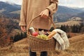 Woman holding wicker picnic basket with thermos, snacks and plaid in mountains on autumn day, closeup Royalty Free Stock Photo