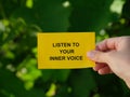 A woman holding a yellow paper note with the words Listen To Your Inner Voice on it against a background of foliage