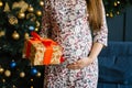 Woman holds hands on her pregnant belly on the background of Christmas lights of fir tree and holds a gift Royalty Free Stock Photo