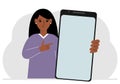 A woman holds a mock-up of a large smartphone with a blank screen and with his second hand points to the screen of the