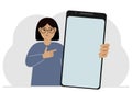 A woman holds a mock-up of a large smartphone with a blank screen and with his second hand points to the screen of the