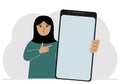 A woman holds a mock-up of a large smartphone with a blank screen and with his second hand points to the screen of the