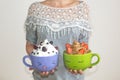 A woman holds two large mugs with painted faces, with whipped cream and berries. Chocolate mousse with strawberries and vanilla