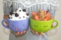 A woman holds two large mugs with painted faces, with whipped cream and berries. Chocolate mousse with strawberries and vanilla