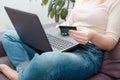 A woman at home sitting on a sofa holds a credit card in her hands and makes a payment using a laptop. happy online shopper