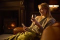 Woman At Home Sitting On Sofa In Lounge With Cosy Fire With Blank Screen Mobile Phone Holding Glass Of Wine Royalty Free Stock Photo