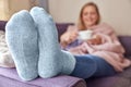 Woman At Home Wearing Cosy Warm Socks And Wrapped In Blanket Lying On Sofa Drinking Cup Of Tea