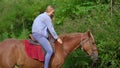 A woman on a horseride in a forest. Her horse is eating plants