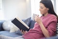 Woman With Hot Drink Relaxing On Sofa At Home Reading Book Royalty Free Stock Photo