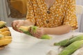 Woman husking corn cob at white wooden table, closeup