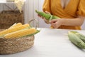 Woman husking corn cob at white wooden table, focus on basket