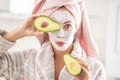 Woman ina dressing gown and towel on her hair wears white face mask and halved avocado over her eye