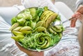 Woman in jeans holding fresh healthy greeen salad with avocado, kiwi, apple, cucumber, pear, greens and sesame on light background