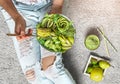 Woman in jeans holding fresh healthy greeen salad with avocado, kiwi, apple, cucumber, pear, greens and sesame on light background