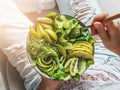 Woman in jeans holding fresh healthy greeen salad with avocado, kiwi, apple, cucumber, pear, greens and sesame on light background Royalty Free Stock Photo