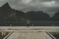 A woman on a jetty facing the sea and the mountains in front of her