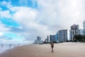 Woman jogs on the beach near skyscrapers of oceanside city with tourists standing in ocean under dramatic cloudscape