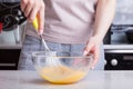 A woman in the kitchen holds a whisk in her hands with a yellow handle and whips them with eggs in a glass transparent bowl. Mothe