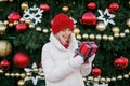 A woman in a knitted red hat with a surprised look holds a gift box in her hands while standing at a Christmas tree in the city sq Royalty Free Stock Photo