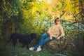 Woman in knitted sweater with short haircut is sitting on fallen tree and black rottweiler dog runs around in the forest