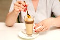 Woman with the latte. Glass mug of latte coffee on the white saucer with female hands holding teaspoon on the table in