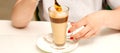 Woman with the latte. Glass mug of latte coffee on the white saucer with female hands holding teaspoon on the table in