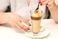 Woman with the latte. Glass mug of latte coffee on the white saucer with female hands holding teaspoon on the table in